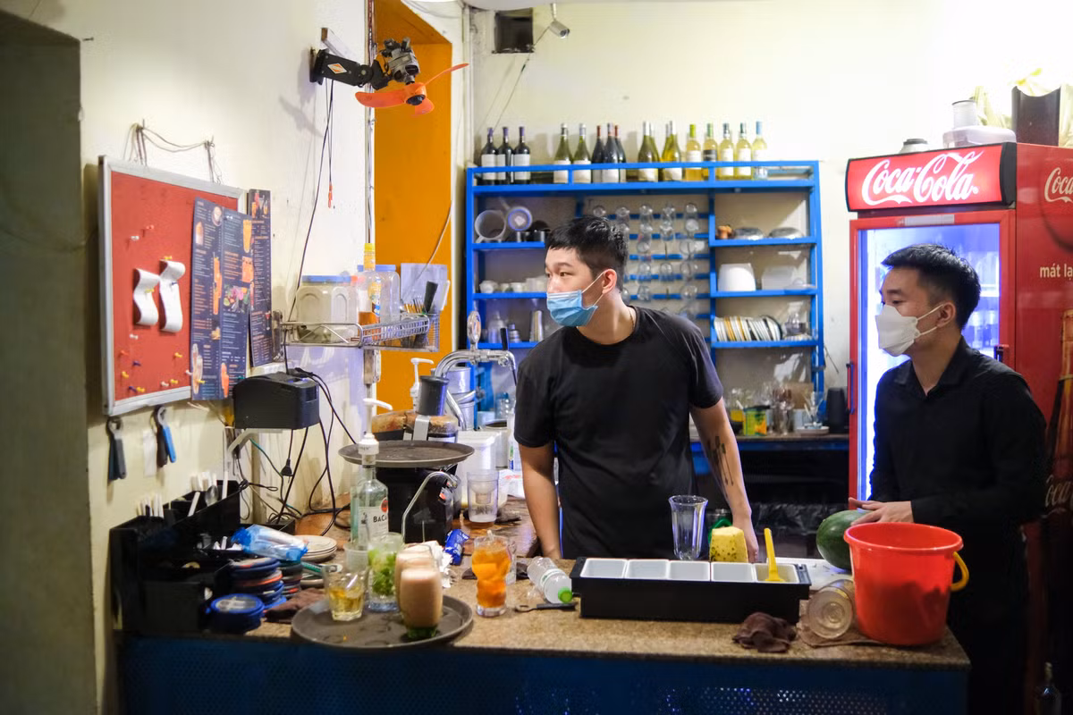 Trong quầy pha chế, Đạt (bên phải) cũng tất bật, liên tục hoạt động không ngơi tay. "Bình thường, mình có thêm một đồng đội nữa, hai người hỗ trợ lẫn nhau. Nay chỉ mình đứng quầy nên phần việc tăng lên gấp đôi", Đạt chia sẻ. Lâu ngày mới động vào cốc chén, dụng cụ pha chế quen thuộc, anh cảm thấy "hơi bị ngợp", "chưa quen tay hẳn".