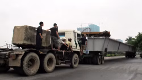 Một xe kéo, một xe đẩy chạy trên đường, công nhân đu bám bên khối bê tông lớn.