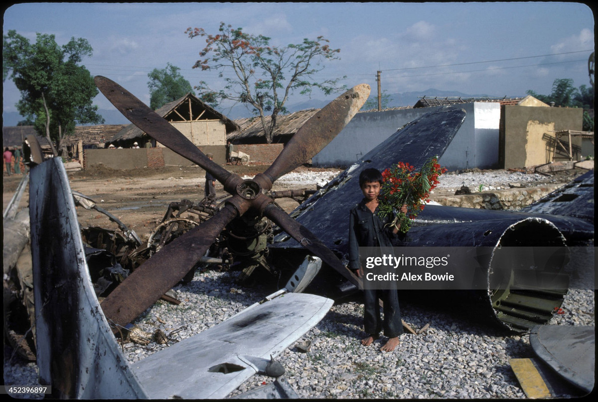 Cậu bé cầm bó hoa phượng đứng bên xác máy bay Pháp trước buổi lễ kỷ niệm 30 năm chiến thắng Điện Biên Phủ, 7/5/1984.