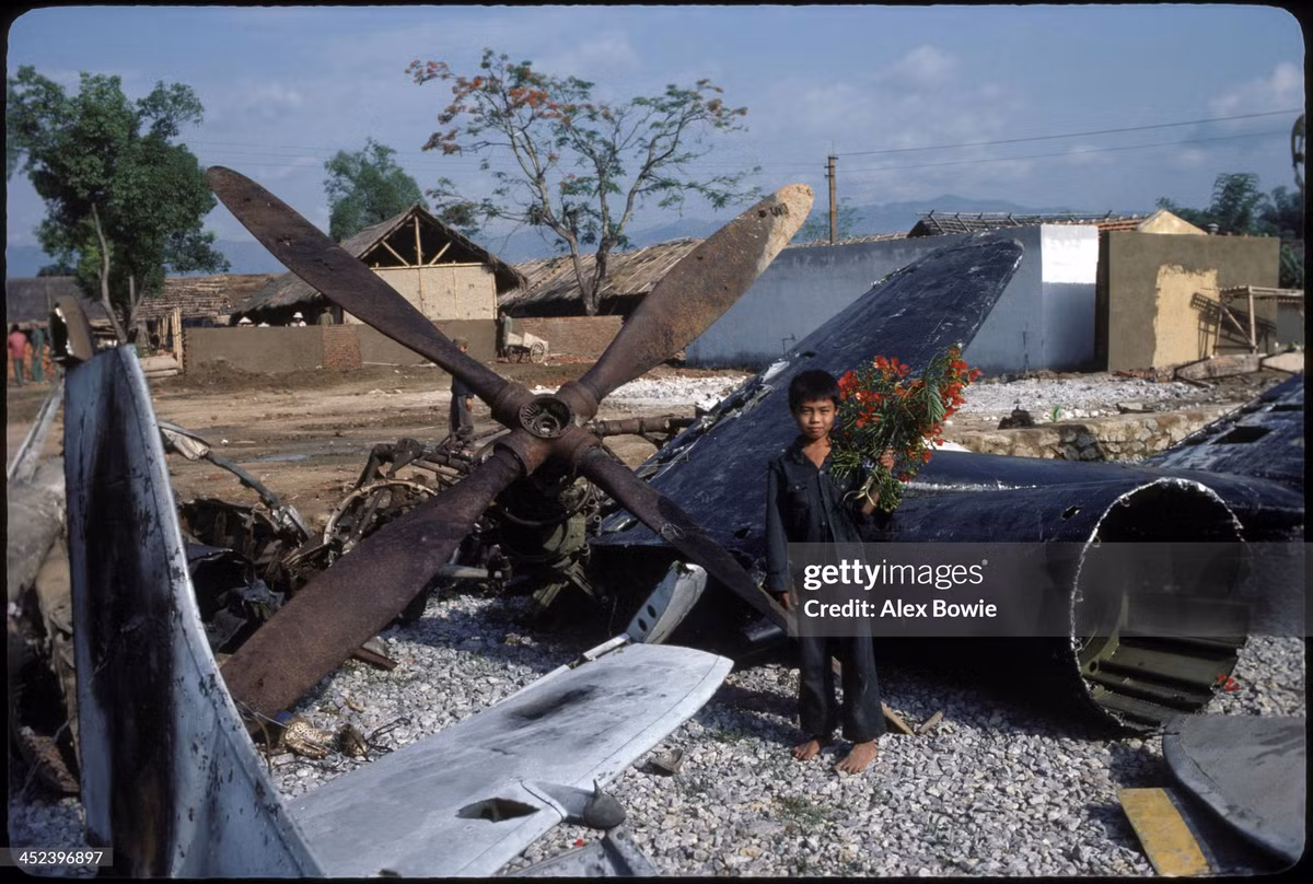 Cậu bé cầm bó hoa phượng đứng bên xác máy bay Pháp trước buổi lễ kỷ niệm 30 năm chiến thắng Điện Biên Phủ, 7/5/1984.