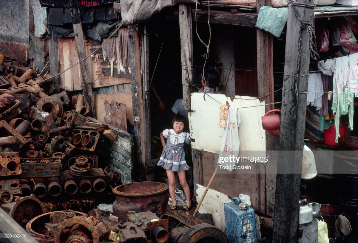 Bé gái đứng bên cửa ngôi nhà lụp xụp. Sân nhà chất đầy phế liệu từ các máy cơ khí cũ.