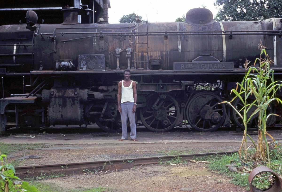Người trông coi nhà ga Enugu đứng bên một đầu máy hơi nước cũ. Đầu máy này thuộc lớp River, một mặt hàng xuất khẩu của Anh.