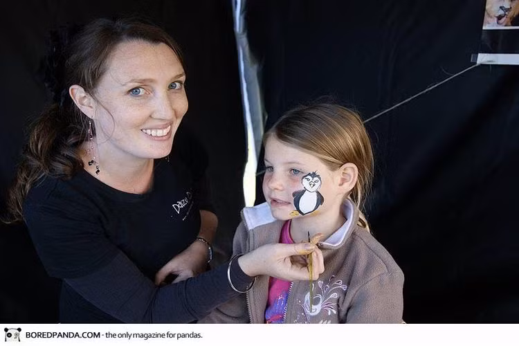 Christy Lewis đang sử dụng màu sơn để tô vẽ lên gương mặt của cô con gái một chú chim cánh cụt.