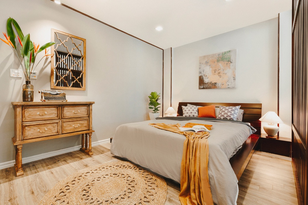 Căn hộ tầng cao được thiết kế theo phong cách Đông Dương kết hợp rustic mộc mạc. Tranh sơn dầu trên vách gỗ vừa là điểm nhấn vừa là vách ngăn không gian ngủ. Cạnh giường là tủ console chân con và gương chia ô hình quả trám cách điệu độc đáo.