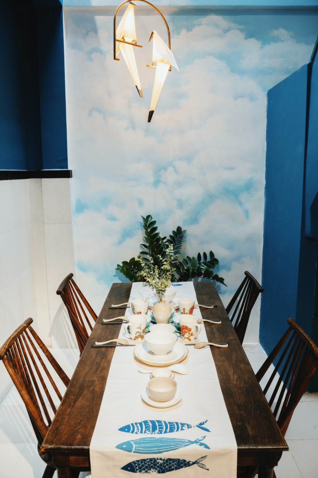 Phòng bếp và khu vực ăn uống được bố trí bàn ghế gỗ tự nhiên cùng điểm nhấn là đôi chim én vàng kết hợp với nền tường họa tiết mây trời. Ngoài ra, khu vực này còn được bố trí giếng trời cùng một chậu cây kim tiền nhằm tạo thêm sinh khí.