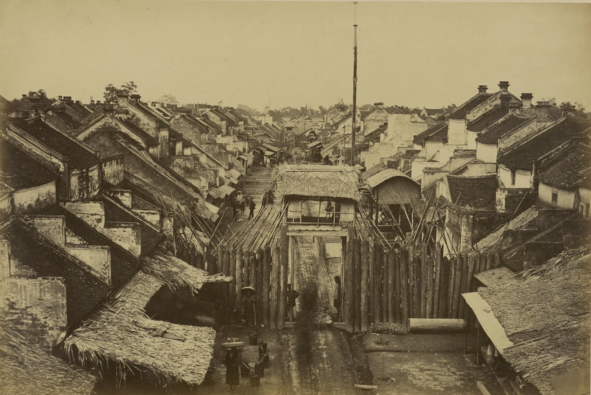 Phố phường Hà Nội. Ở tiền cảnh của bức ảnh, chúng ta có thể nhìn thấy cánh cổng đóng kín đường phố. Hầu hết đường phố và khu phố của Hà Nội được ngăn cách bởi những cánh cổng lớn được đóng vào ban đêm, được canh gác bởi những người canh gác ở chòi canh phía trên cổng.