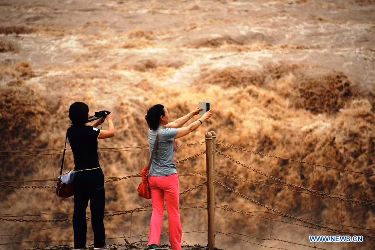 2 khách du lịch mải mê ghi lại hình ảnh dòng nước lũ cuồn cuộn, đục ngầu phù sa.