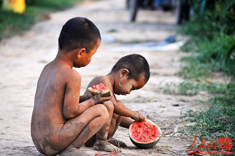 Hai đứa trẻ người lấm lem bùn đất chia nhau miếng dưa hấu.