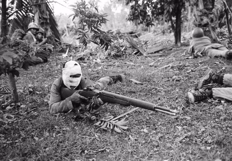 Một binh sĩ Sài Gòn bị thương và phải băng bó khắp đầu, chỉ hở đôi mắt và miệng nhưng vẫn cầm khẩu súng trên tay trong tư thế sẵn sàng chiến đấu với bộ đội Việt Nam ở khu vực Đồng Xoài ngày 11/6/1965.