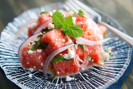 Salad dưa hấu. Nguyên liệu: 1 /2 quả dưa hấu, 1 quả hành tây, ít lá bạc hà, chanh, dầu olive, 1 ít phô mai vụn. Dưa hấu gọt vỏ, cắt miếng vuông vừa ăn. Hành tây xắt lát mỏng. Nếu sợ hành quá hăng bạn có thể ngâm vào tô nước đá lạnh khoảng 15 phút rồi vớt ra, để ráo. Cho hành và dưa hấu vào tô, thêm phô mai và lá bạc hà xắt nhỏ. Rưới dầu olive và nước chanh vào tô salad, trộn đều.