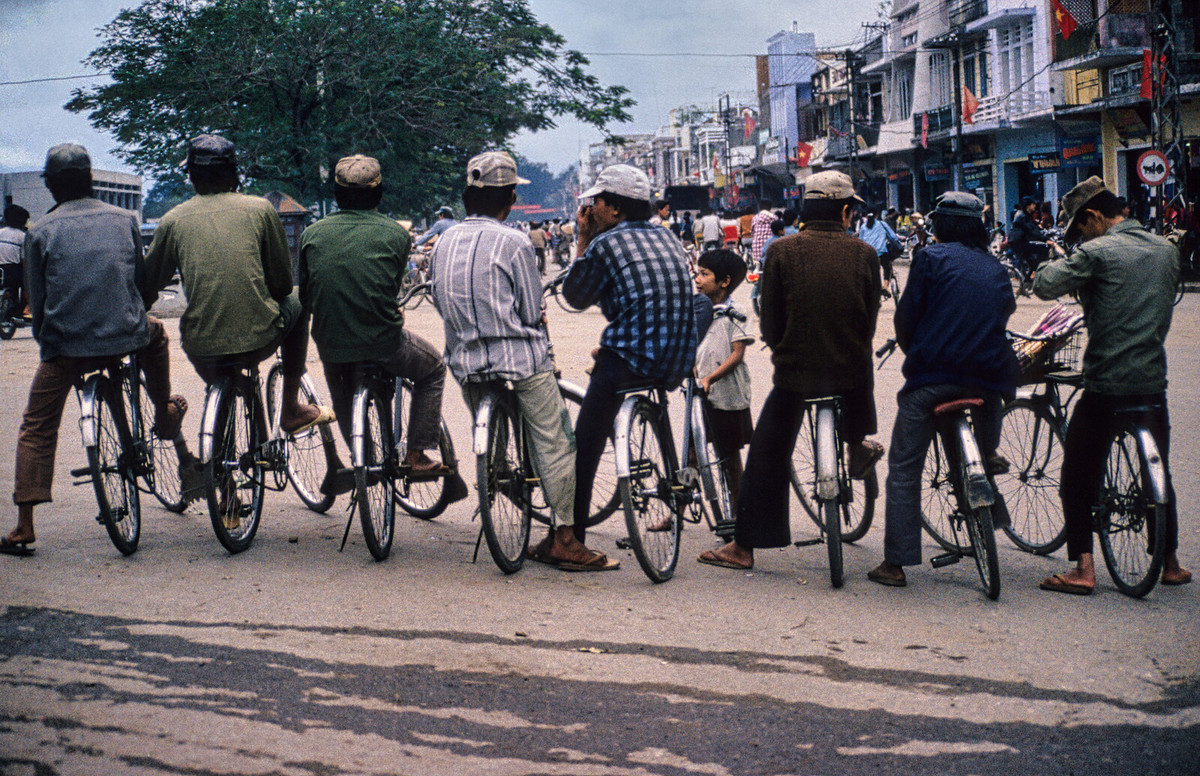 Các nam giới ngồi xe đạp dàn hàng trên đường Trần Hưng Đạo. Chợ Đông Ba phía bên trái.