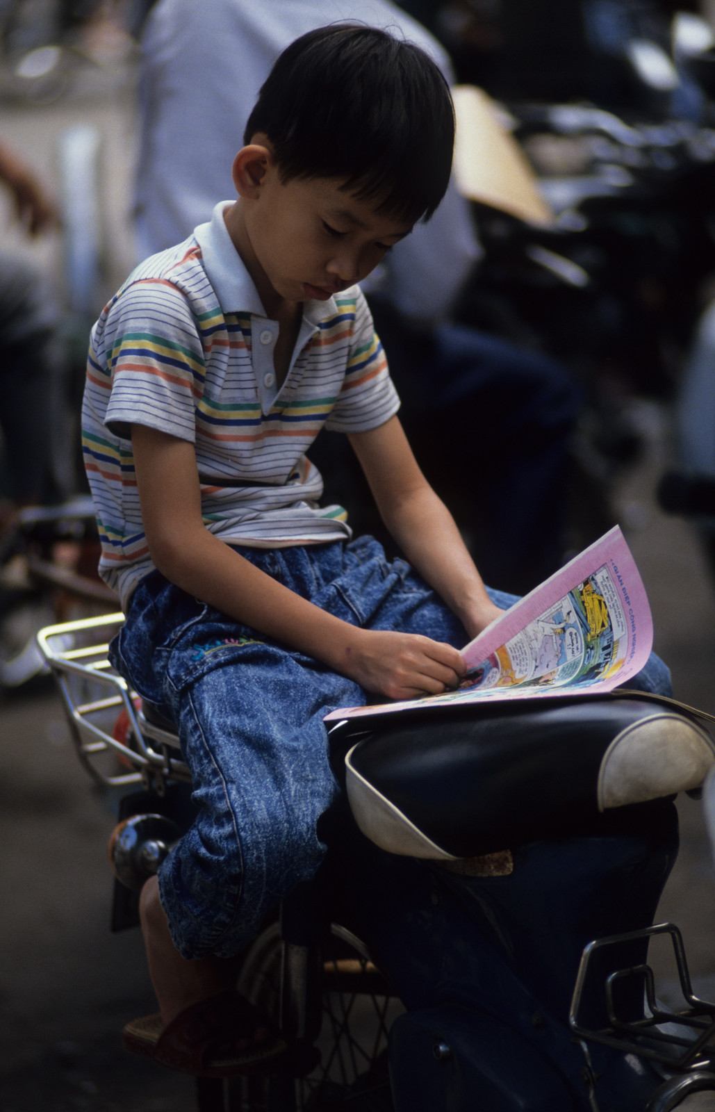 Chú bé ngồi đọc báo trên xe máy.