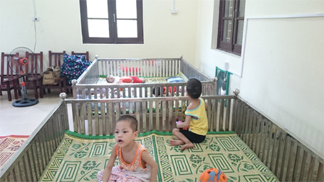 Căn phòng có diện tích rộng khoảng 60 m2, với đầy đủ quạt cây, bàn ăn... Trong ảnh là hai trẻ em chùa Bồ Đề chơi trong cũi sắt do còn khá lạ lẫm khi chuyển đến nơi ở mới.