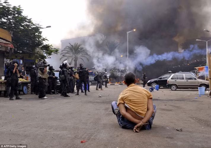 Một người biểu tình Ai Cập bị trói tay ngồi bệt giữa đường, ngay cạnh đó là một nhóm quân đội đang tiếp tục đàn áp những người biểu tình khác.