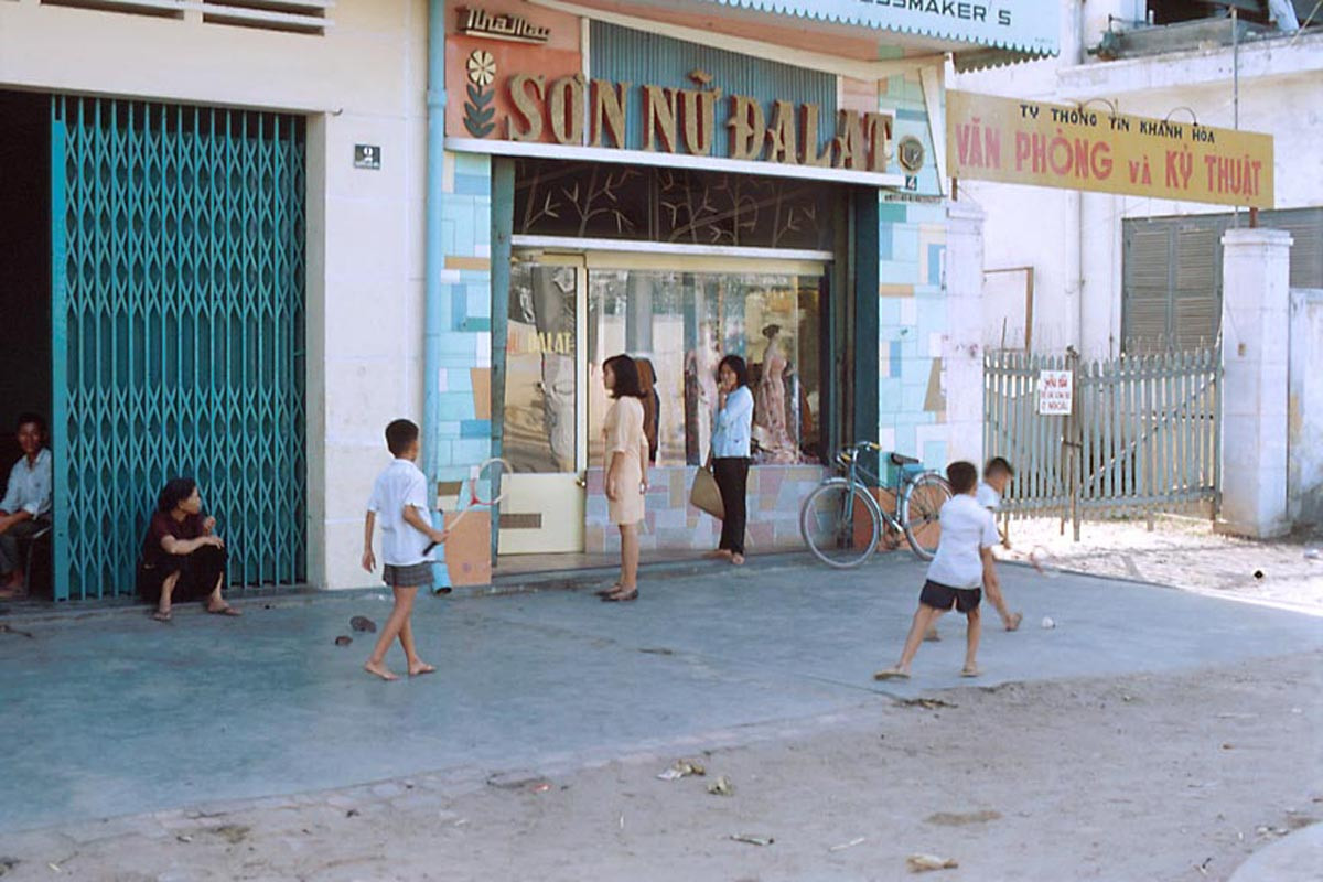Những đứa trẻ chơi cầu lông bên ngoài tiệm may "Sơn nữ Đà Lạt", cạnh đó là cổng vào văn phòng của Ty thông tin Khánh Hòa.
