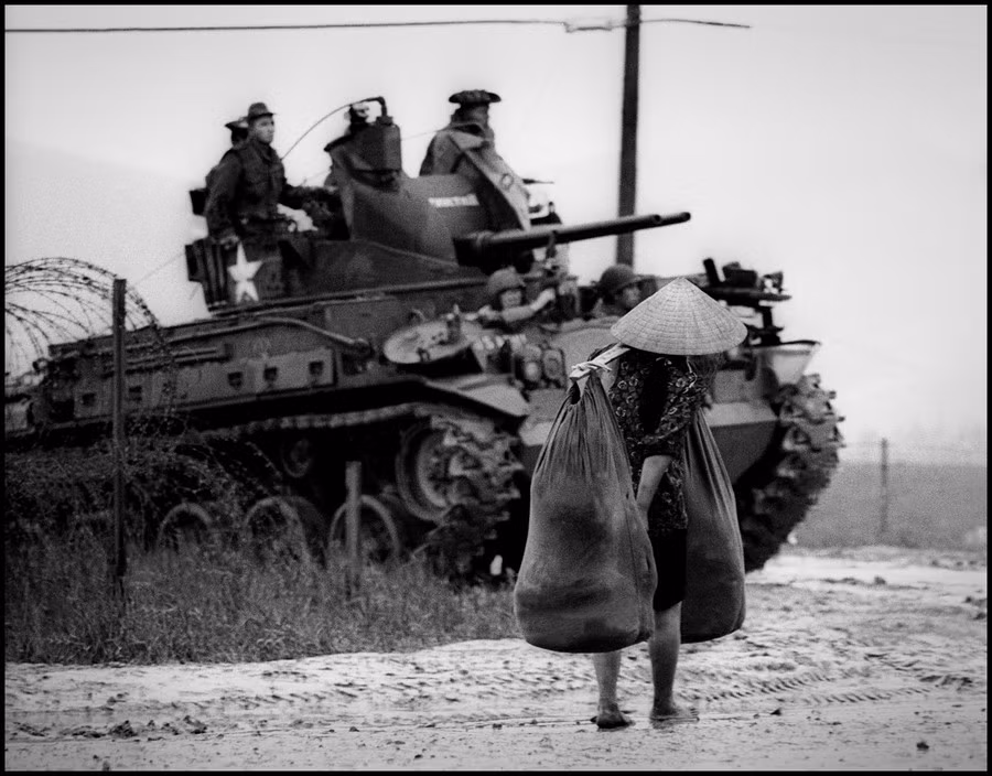 Một người phụ nữ địa phương gánh đồ đạc đi ngang qua một xe tăng Mỹ lúc di tản khỏi vùng chiến sự. Ảnh: Philip Jones Griffiths.