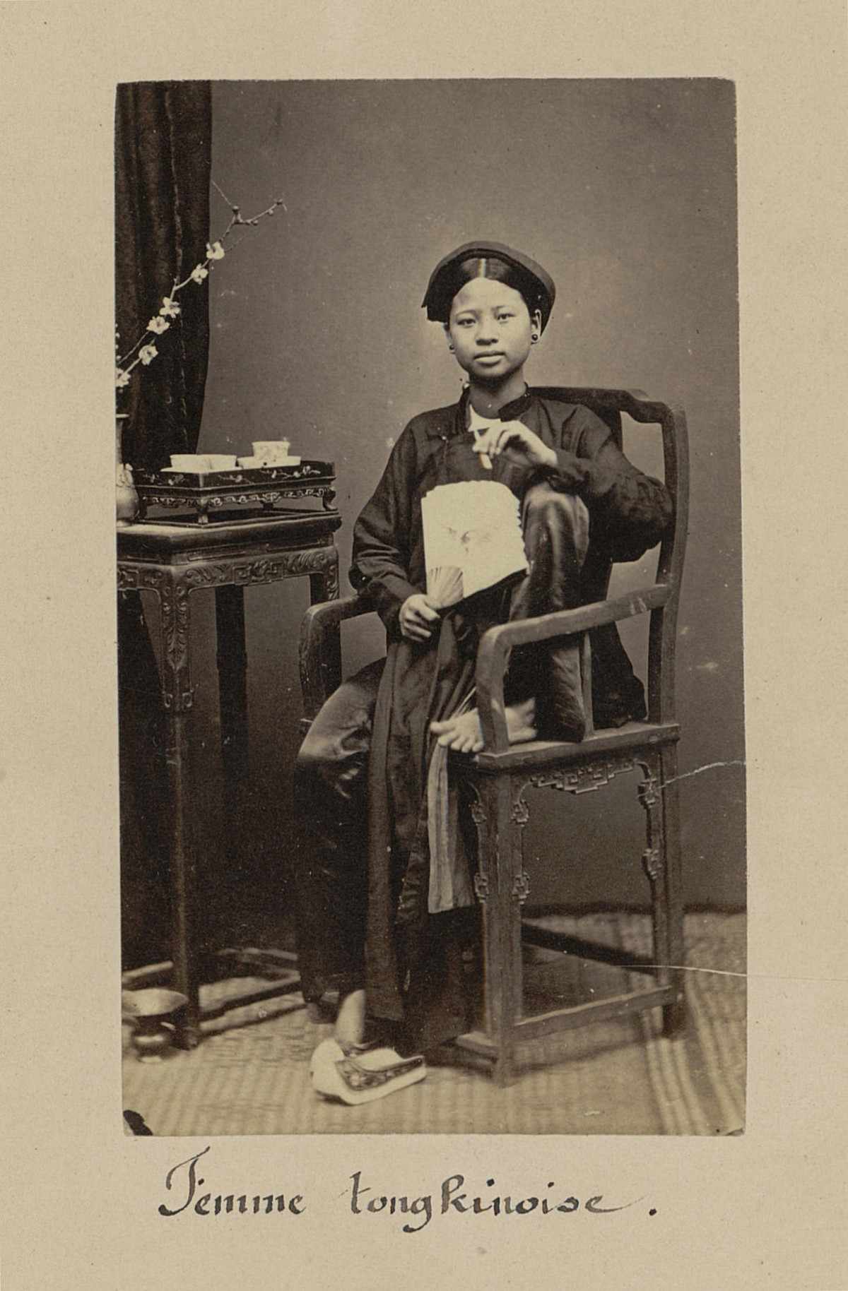 Ảnh thiếu nữ xứ Bắc Kỳ cầm quạt, đầu đội khăn xếp, ngồi trên ghế gỗ.