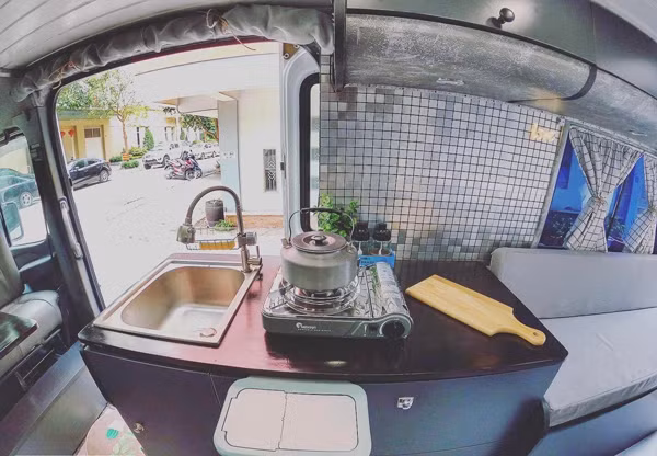 Vì diện tích sử dụng chỉ 6m2 nên phải tận dụng tối đa không gian và các đồ vật trong xe có nhiều công dụng nhất”, anh Sơn chia sẻ. Trong khu bếp, vợ chồng anh Sơn sử dụng một chiếc tủ lạnh chạy nguồn 12V, dung tích 50l giúp bảo quản thức ăn. Và bếp ga mini được sử dụng trong căn bếp này để tiện việc mang ra ngoài nấu nướng khi cần. Bồn rửa tay và hoa quả được đặt đối diện bếp để tiện sử dụng. Dưới bồn là một bình nước 110l để cung cấp nước cho cả xe. Từ bình nước này, đôi vợ chồng trẻ thiết kế một đường nước dẫn tới vòi xịt cũng như vòi hoa sen ở đằng sau. Đường điện được bố trí phía đối diện và tách biệt với đường nước.