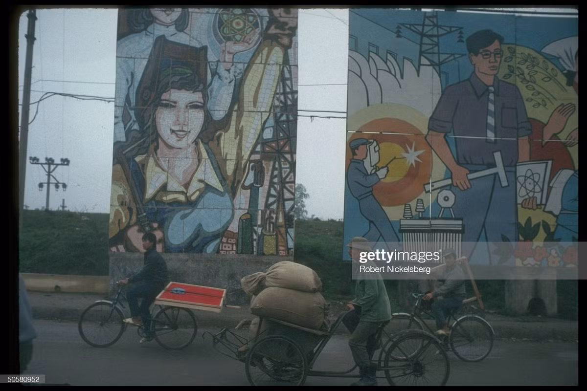 Xe đạp và xích lô chở hàng ngang qua những bức tranh cổ động.