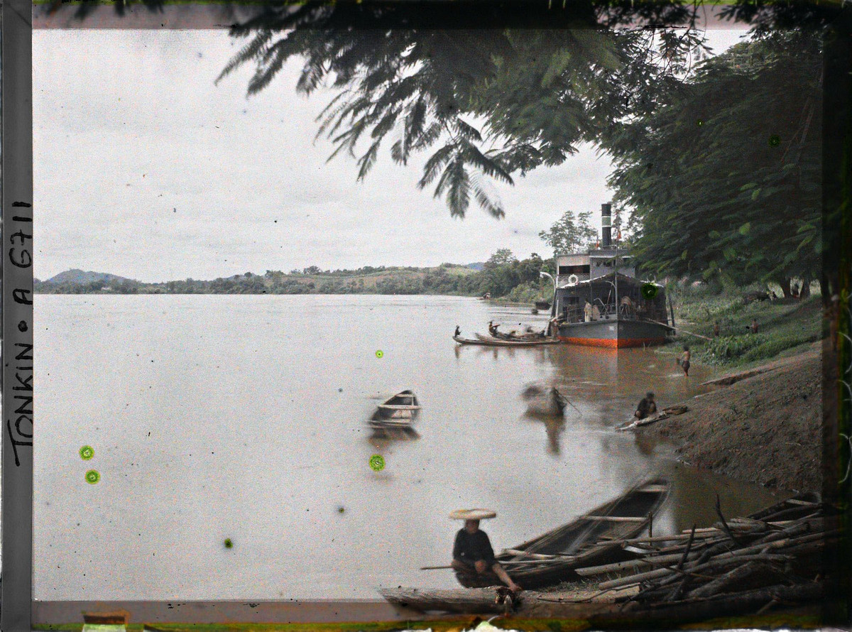 Tàu hơi nước của Pháp neo đậu bên bờ sông Lô ở phủ Đoan Hùng. Dân địa phương chèo thuyền tam bản đến gần để chào bán sản phẩm địa phương.