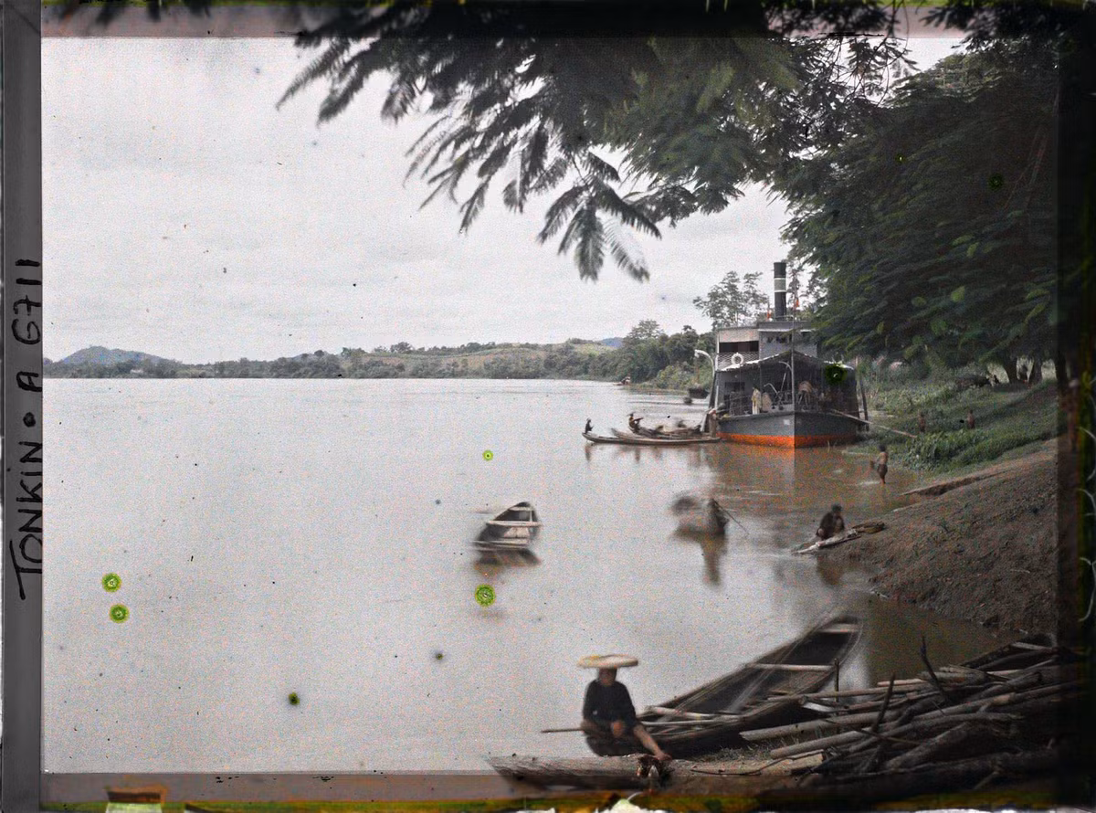 Tàu hơi nước của Pháp neo đậu bên bờ sông Lô ở phủ Đoan Hùng. Dân địa phương chèo thuyền tam bản đến gần để chào bán sản phẩm địa phương.