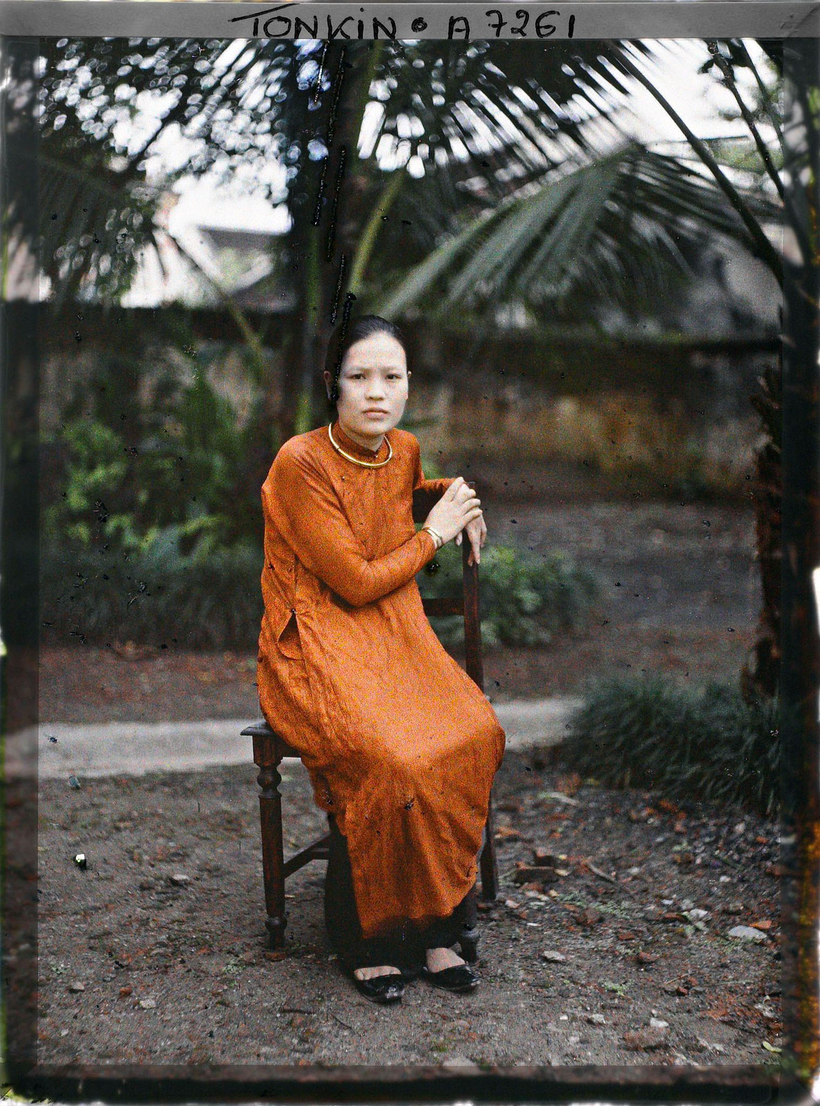 Cô gái trẻ mặc áo dài màu cam, cổ đeo kiềng vàng, ngồi trên ghế gỗ.