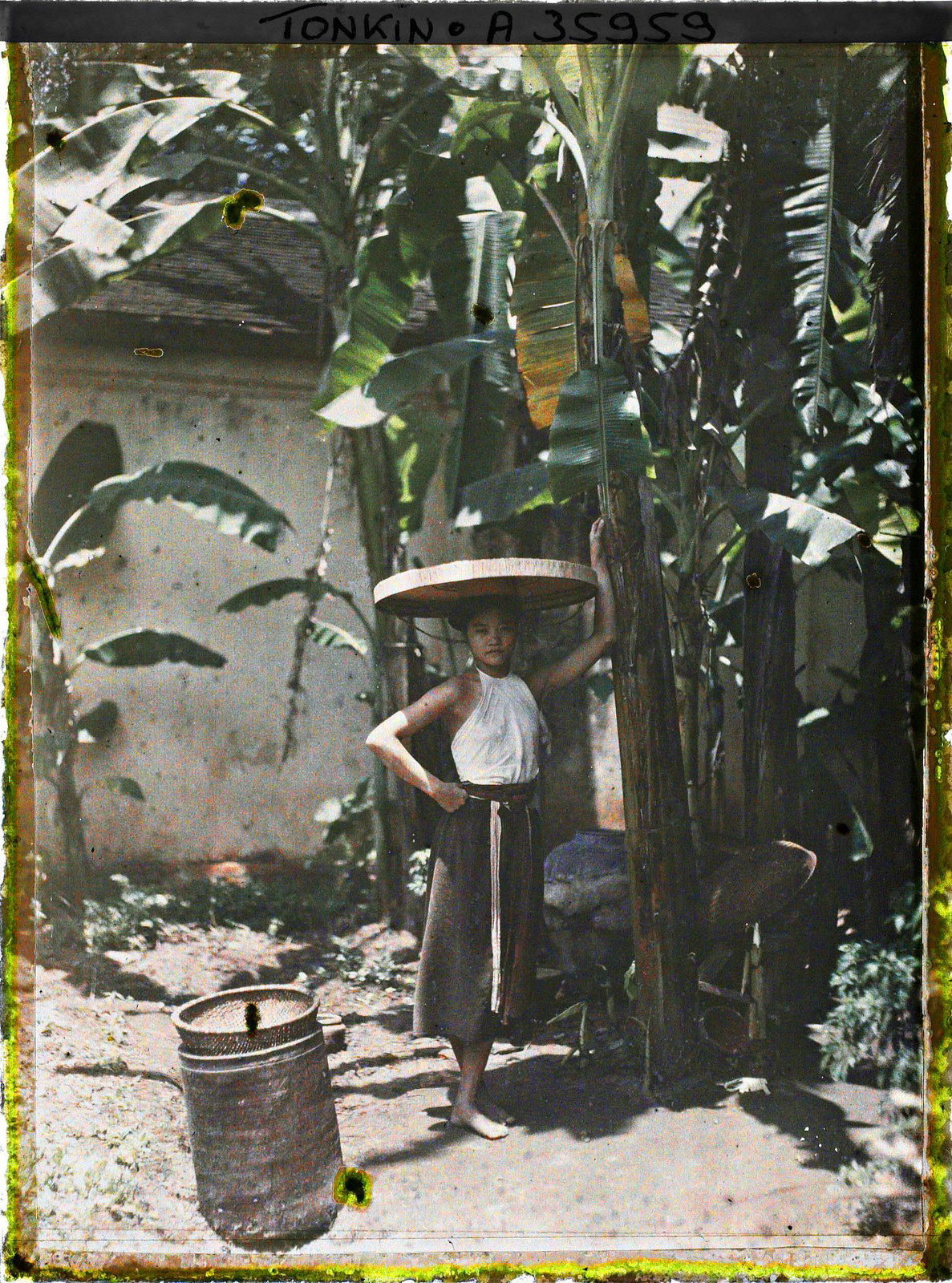 Cô thôn nữ mặc áo yếm, đội nón quai thao, tạo dáng bên cây chuối, Bắc Bộ năm 1921. Ảnh: Léon Busy/Collections.albert-kahn.hauts-de-seine.fr.