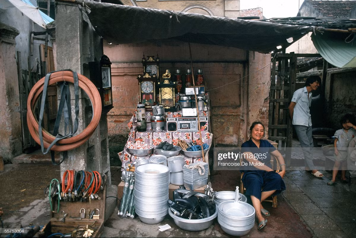 Một cửa hàng trên vỉa hè phố cổ. Hàng hóa được bày bán gồm nồi, khay đá và chậu nhôm, đồng hồ để bàn, đài cát-xét, phích Rạng Đông, phích đá Liên Xô, bơm xe đạp, lốp và yên xe đạp, các loại khóa...