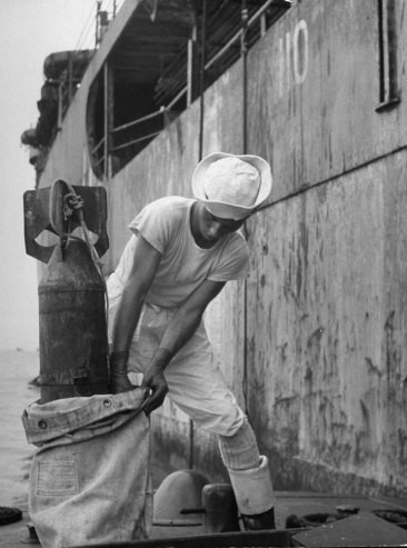 Nhân viên Mỹ lấy một quả bom mẫu chứa vi khuẩn ở trong bao tải để kiểm tra bức xạ sau khi đó được hạ xuống từ tàu sân bay USS Bracken (nền) trong quá trình thử nghiệm bom hạt nhân dưới nước ở đảo san hô Bikini Atoll, thuộc quần đảo Marshall ở Thái Bình Dương.