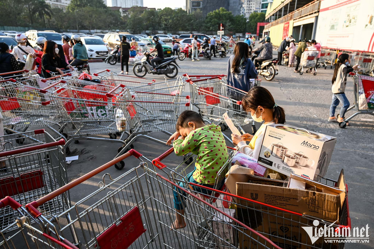 Phía ngoài cổng, nhiều xe đẩy hàng được bỏ lại sau khi khách đã ra về. Lượng người đổ về đông khiến cho việc lấy ô tô, xe máy cũng mất thêm nhiều thời gian. Trong hình, hai em nhỏ chờ cha mẹ bên giỏ đầy ắp hàng Tết.