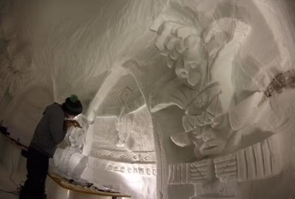 Một thợ điêu khắc đang tạc một bức tượng từ một phiến băng bên trong một lều tuyết.