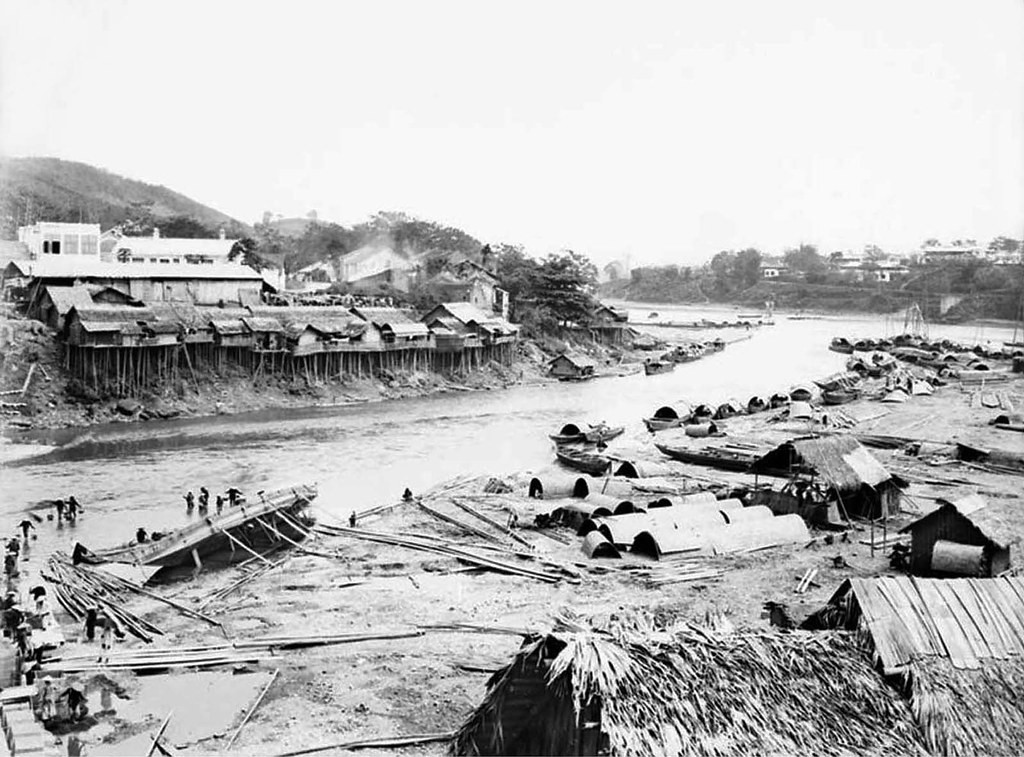 Thị trấn Lào Cai nhìn từ đầu phía Trung Quốc của cầu Nậm Thi. Để chống lại dòng nước chảy siết, những ngôi nhà được xây dựng trên sườn đồi và được chống đỡ bằng những cọc tre.