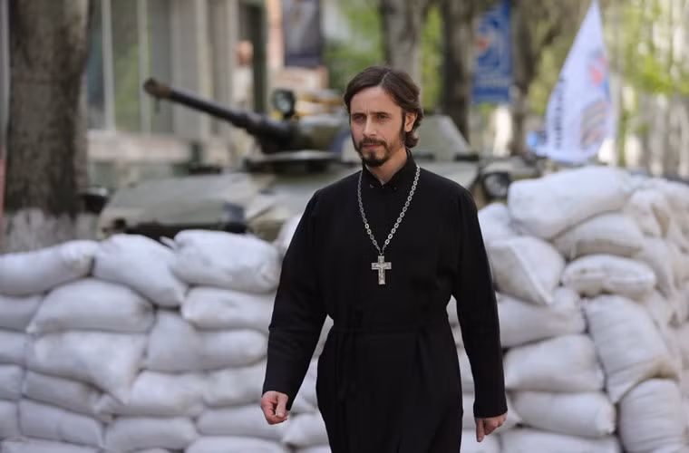 Một tu sĩ đi qua các công sự ở Slavyansk, đằng sau ông này xe chiến đấu lính dù BMD-1 đặt trong công sự bằng bao cát.