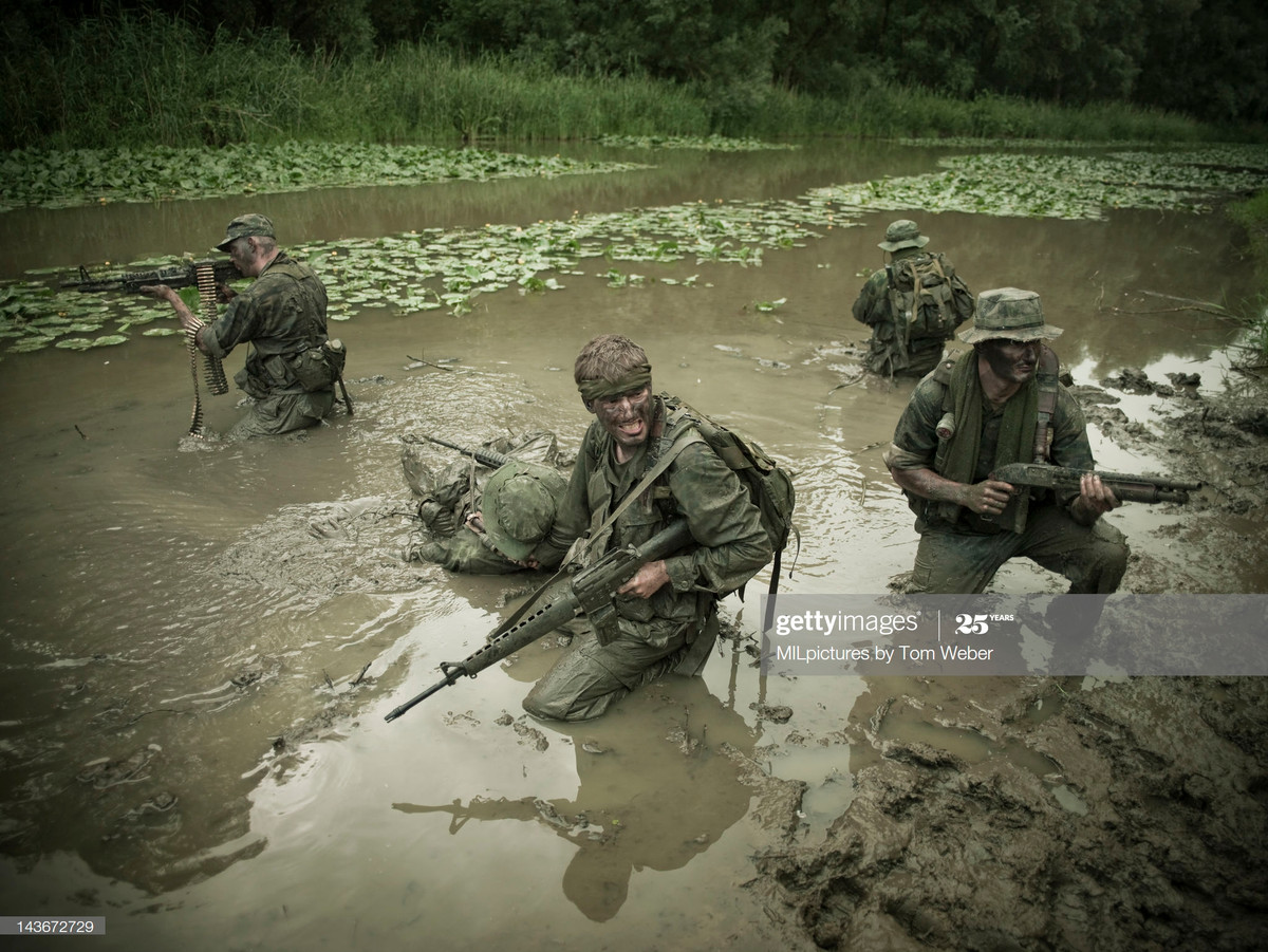 Một nhóm lính Mỹ chĩa súng về bốn phía trong khi một lính Mỹ khác bị thương đang được kéo đi trong bùn lầy, ảnh dàn dựng về cuộc chiến tranh Việt Nam. Ảnh: Tom Weber / MIL Pictures / Getty Images.