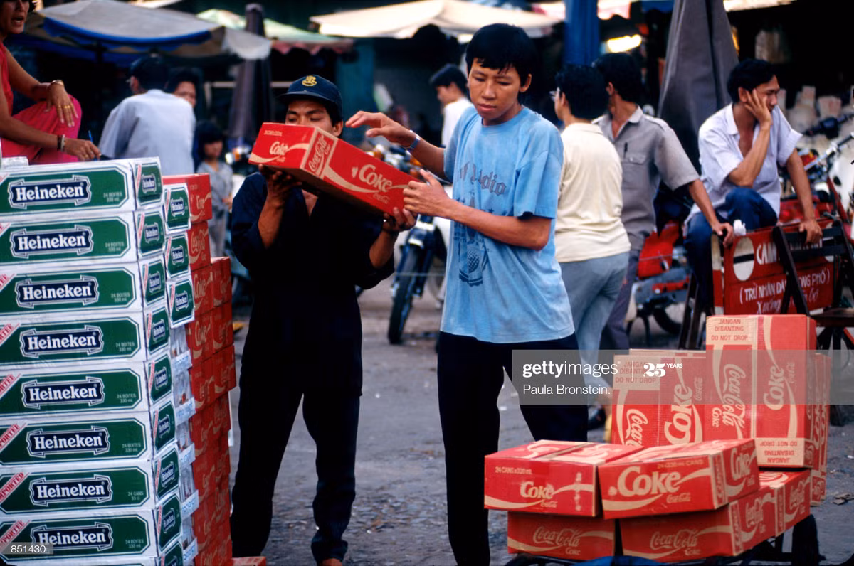 Hai nam giới bốc dỡ các thùng bia Heineken và nước ngọt Coca-Cola tại một khu chợ ở TP HCM, tháng 6/1993. Tại Việt Nam năm 1993, các sản phẩm tiêu dùng phương Tây đã trở nên phổ biến. Ảnh: Paula Bronstein/Getty Images.