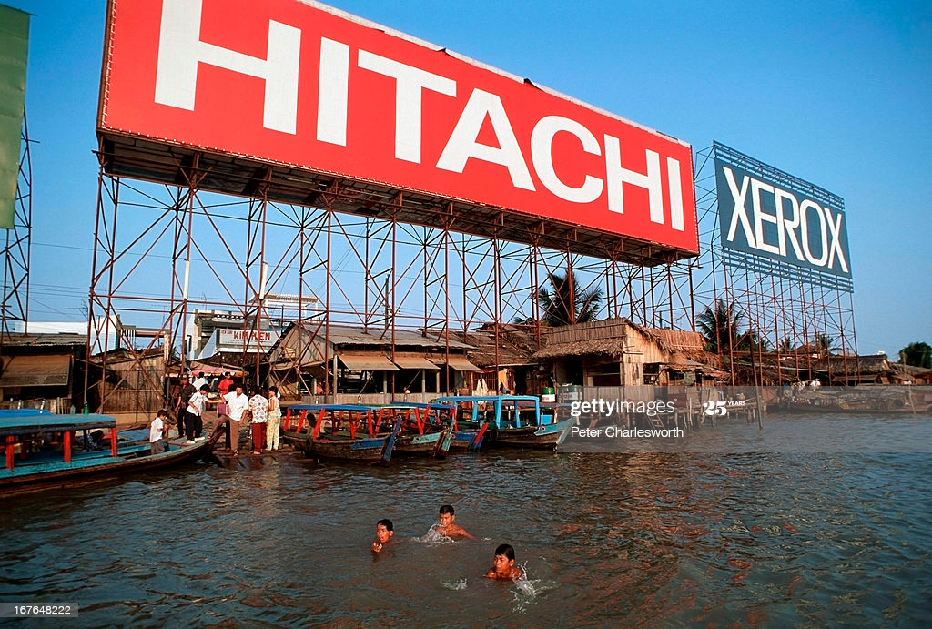 Trẻ em bơi lội ở sông Sài Gòn, gần bến đò trên bán đảo Thủ Thiêm. Trên bờ sông là những ngôi nhà tạm bợ và pa-no quảng cáo khổng lồ.