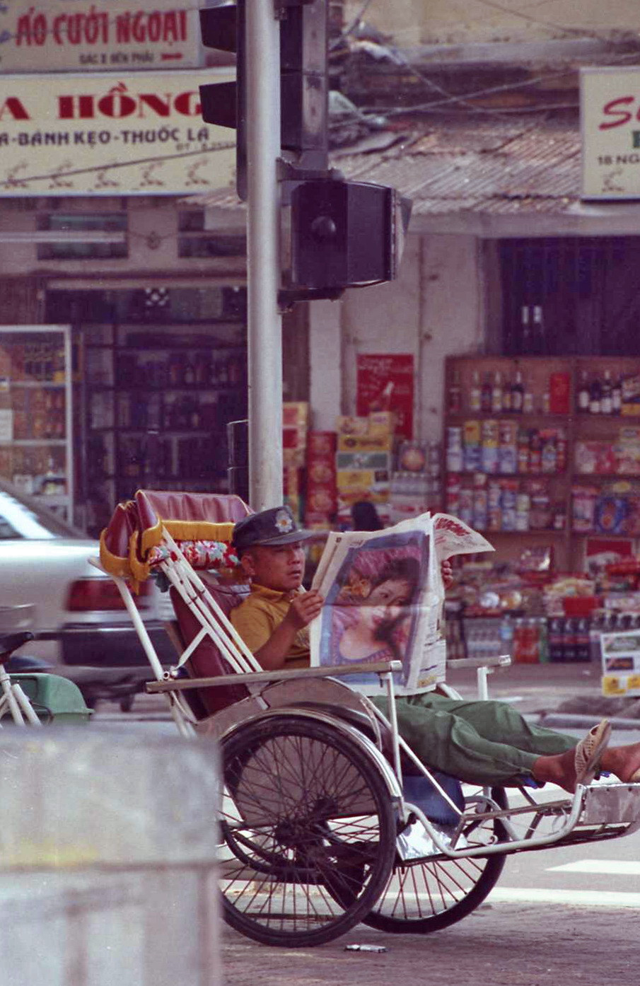 Người lái xích lô đọc báo khi nghỉ ngơi trên phố. Bên kia đường là các sạp bánh kẹo, rượu bia nhập ngoại. Hình chụp năm 1997, trêm con phố có tên bắt đầu bằng chữ "Ng" (Ngõ Gạch?). Ảnh: Yuichi Kobayashi Facebook.