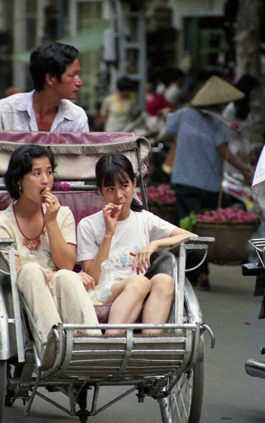 Hai thiếu nữ đang ăn gì đó, hình như là những múi mít, khi ngồi trên xích lô vào một chiều chủ nhật nóng nực ở Hà Nội, 1998. Người lái xích lô trông giống tài tử Jean-Paul Belmondo thời trẻ, người chụp ảnh nhận xét. Ảnh: Yuichi Kobayashi Facebook.
