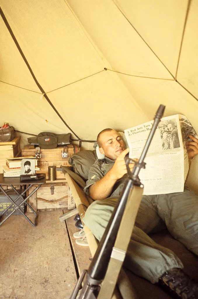 Đọc báo khi nằm trong lều dã chiến với khẩu súng gác bên thành giường. Ảnh: Jean-Claude Sauer.