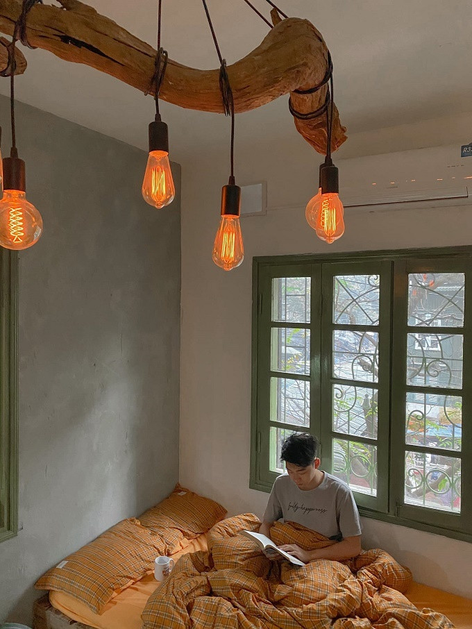 Toàn bộ nội thất như: Giường ngủ, bàn học, bàn ăn, kệ tủ, tủ quần áo...Nhật tự đóng nên chỉ mất tiền mua gỗ. Sau khi hoàn thiện, căn hộ có đầy đủ công năng, thoáng đãng và đẹp như homestay. Ảnh: Giường ngủ bên khung cửa sổ, đón ánh nắng tự nhiên. Mỗi khi rảnh rỗi, Nhật nghỉ ngơi, đọc sách. Đèn chân nhện được anh mua trên một trang mạng điện tử, còn cây gỗ mua trên hội gỗ lũa nhìn rất ấm áp. Loại đèn dây tóc edison này hơi tốn điện nhưng thay quạt sưởi vào mùa lạnh, chỉ cần bật 15 phút là ấm phòng. Mùa hè nóng nực có thể thay bằng đèn led.