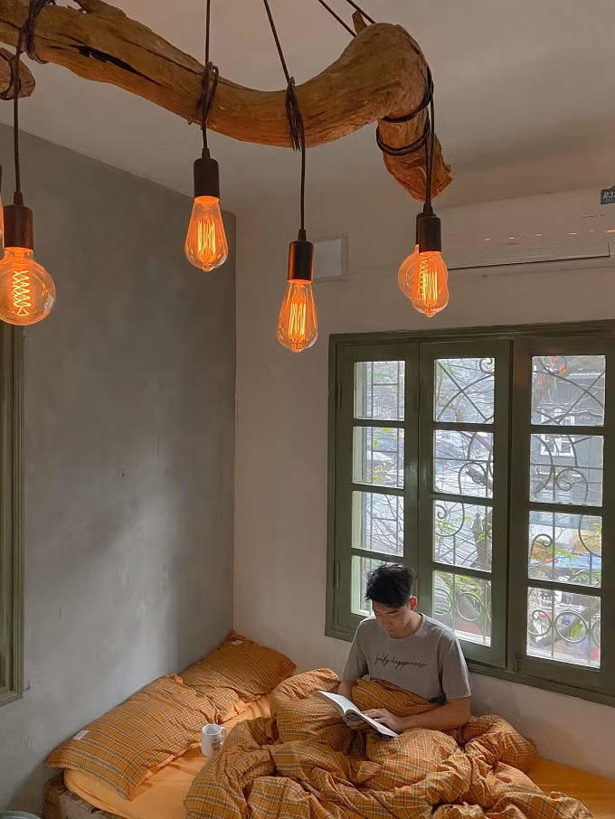 Toàn bộ nội thất như: Giường ngủ, bàn học, bàn ăn, kệ tủ, tủ quần áo...Nhật tự đóng nên chỉ mất tiền mua gỗ. Sau khi hoàn thiện, căn hộ có đầy đủ công năng, thoáng đãng và đẹp như homestay. Ảnh: Giường ngủ bên khung cửa sổ, đón ánh nắng tự nhiên. Mỗi khi rảnh rỗi, Nhật nghỉ ngơi, đọc sách. Đèn chân nhện được anh mua trên một trang mạng điện tử, còn cây gỗ mua trên hội gỗ lũa nhìn rất ấm áp. Loại đèn dây tóc edison này hơi tốn điện nhưng thay quạt sưởi vào mùa lạnh, chỉ cần bật 15 phút là ấm phòng. Mùa hè nóng nực có thể thay bằng đèn led.