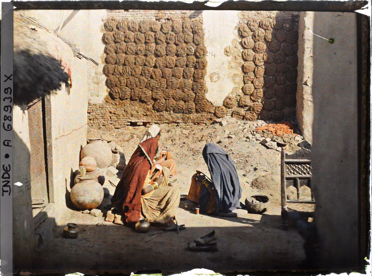 Một nhóm phụ nữ đang đóng giày ở sân nhà, cạnh bức tường được dùng để phơi những miếng phân bò làm chất đốt.