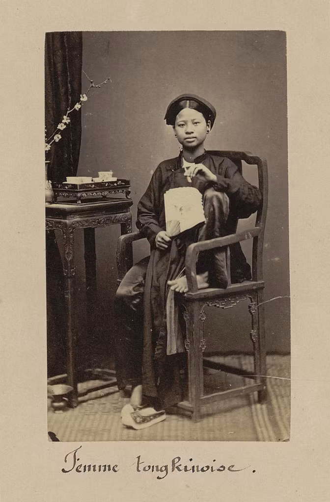 Thiếu nữ xứ Bắc Kỳ cầm quạt, đầu đội khăn xếp, ngồi trên ghế gỗ.