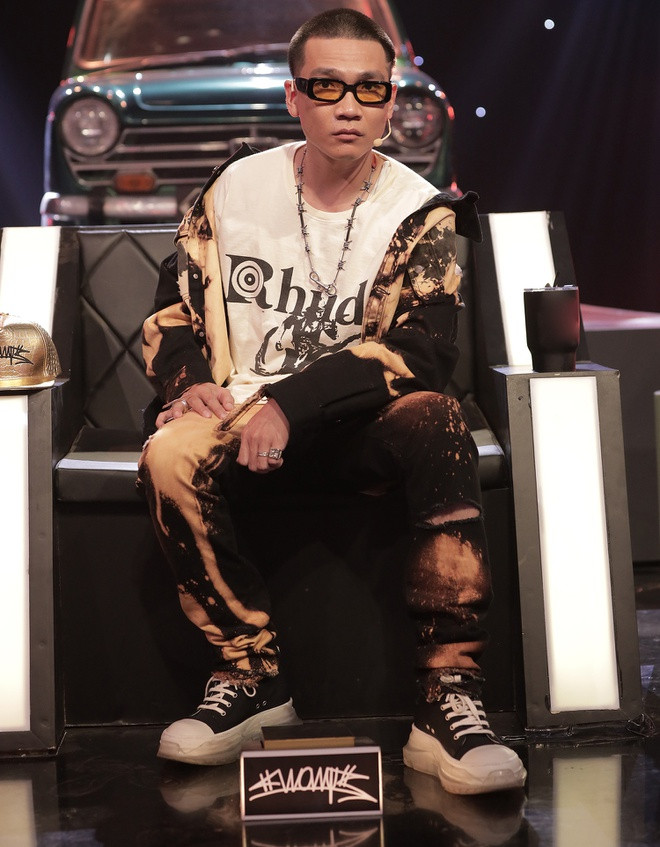 Stylist Ryan Sơn Hoàng cho rằng nam huấn luyện viên muốn thử nghiệm nhiều trang phục khác lạ để lột tả tinh thần đường phố bụi phủi cũng như tính cách của bản thân. Trong tập 10 của Rap Việt, Wowy mặc bộ cánh cá tinh in họa tiết loang lổ màu sắc cùng áo thun trắng của Rhude có giá 249 USD và vòng cổ kẽm gai độc đáo. Ảnh: Rap Việt.