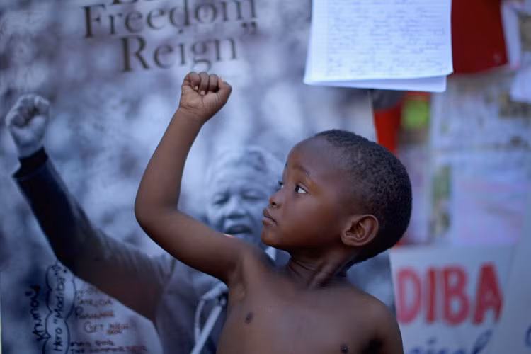 Một bé trai giơ cao nắm đấm, bắt chước hình ảnh chân dung của cựu Tổng thống Nam Phi Nelson Mandela ở bức tưởng tưởng niệm ông bên ngoài Viện tim Medi-Clinic ngày 14/7.