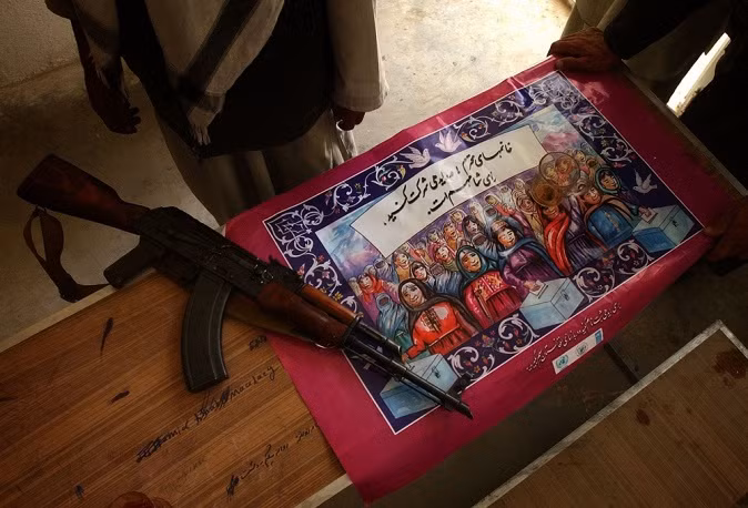 . Khẩu AK đặt trên tấm áp phích cổ vũ người dân tham gia bỏ phiếu ở Khwaja Omri, Afghanistan năm 2004.