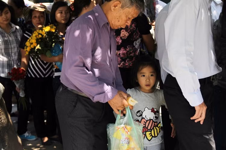 Một em nhỏ xếp hàng vào kính viếng, được ông nội chuẩn bị cả bánh mì theo sau, phòng khi đói bụng để ăn.