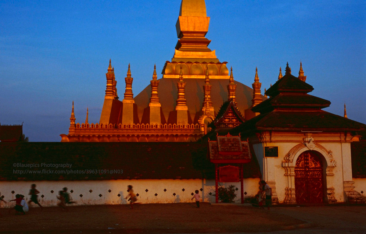 Trẻ em chạy nhảy bên ngoài chùa That Luang lúc hoàng hôn.
