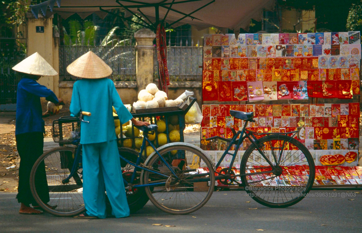 Hàng bán thiệp Tết vỉa hè và chiếc xe bán bưởi bên lề đường.