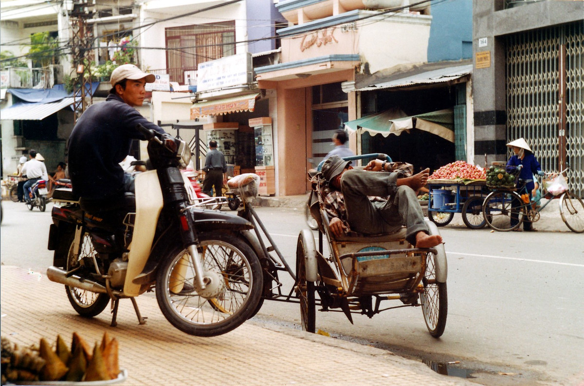 Người lái xe ôm đợi khách cạnh một người lái xích lô đang ngủ trưa trên chiếc xe của mình.